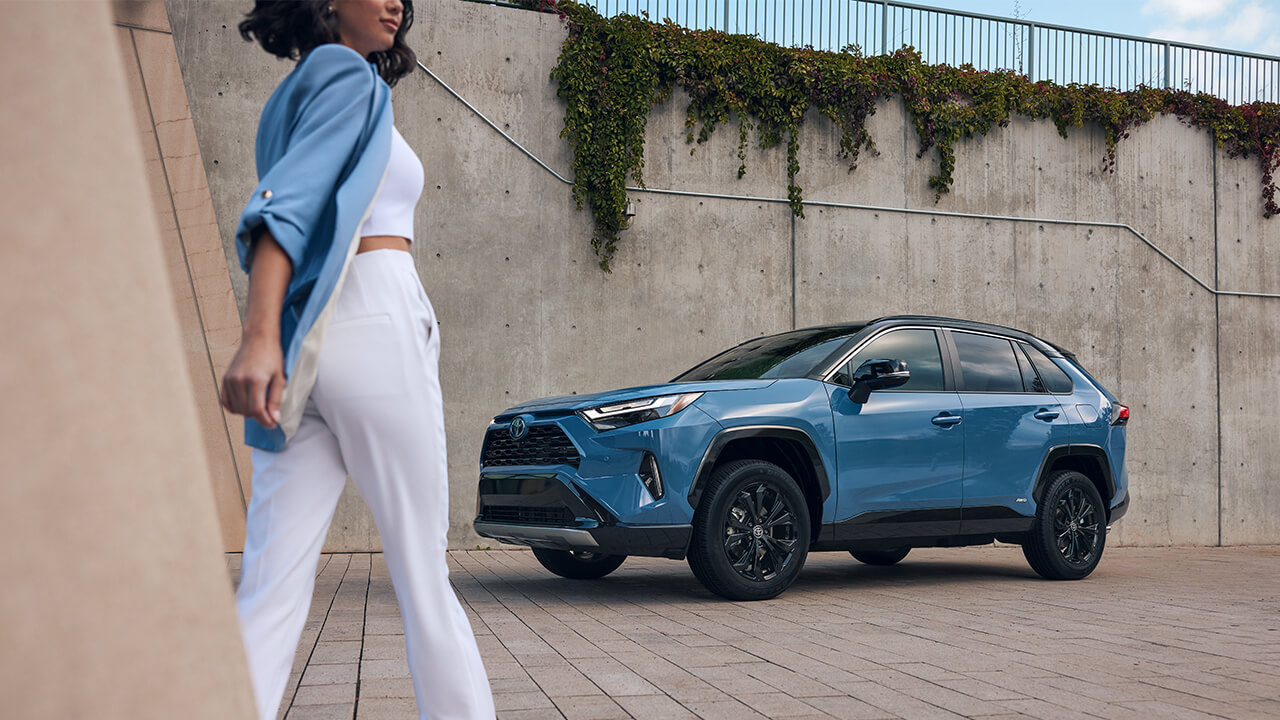 Woman in blue shirt and white plants walking to her Blue 2022  Toyota RAV4 Hybrid
