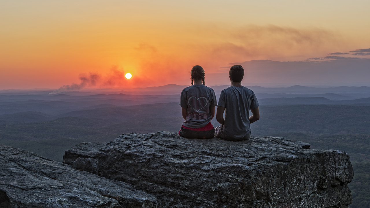 At Alabama's Cheaha Scenic Overlook, a man and a woman sit on a boulder and look over Cheaha Mountain's valleys at sunset.