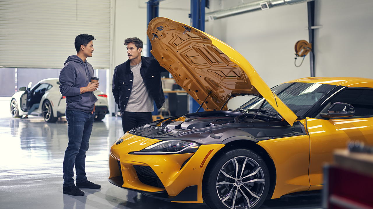 Two men inspect a bright yellow Toyota sports car in an auto garage. 
