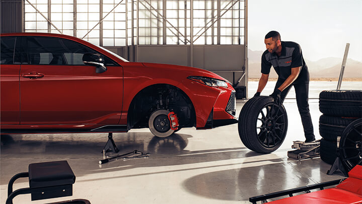 A mechanic rolls a wheel toward a red Toyota vehicle in an open garage. 