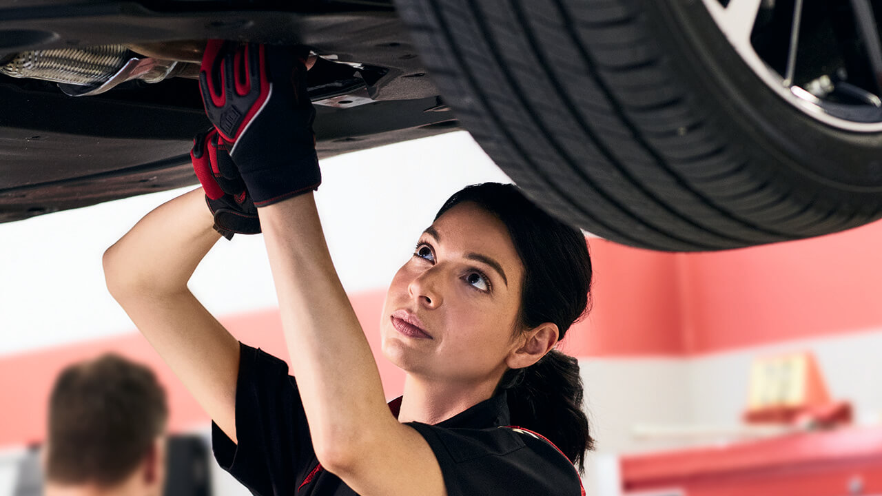 A woman inspects the underside of a car. 