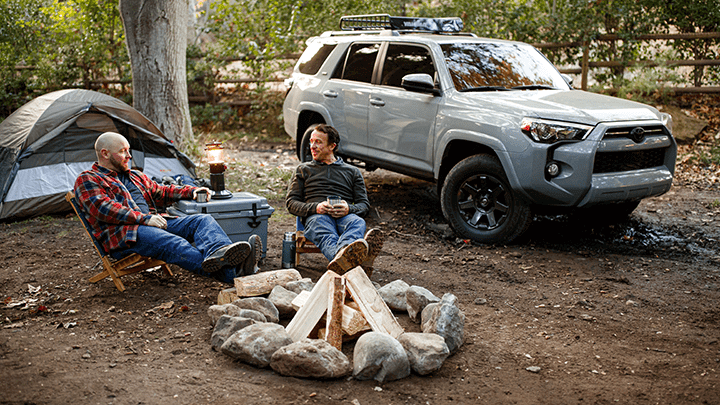 Two men sit by a campfire in the forest, surrounded by a camping tent to the left and a 4Runner to the right.